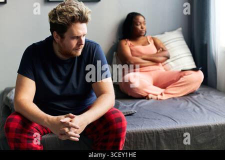 Horizontal shot young Caucasian man and his Black girlfriend sitting on bed in morning in total silence ignoring each other after serious quarrel Stock Photo