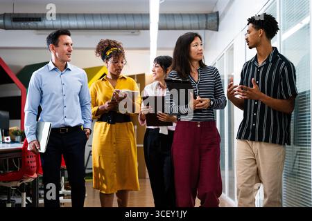 Startup business team members walking through office corridor, holding tablets, laptops, and clipboards, discussing work and collaborating on project Stock Photo