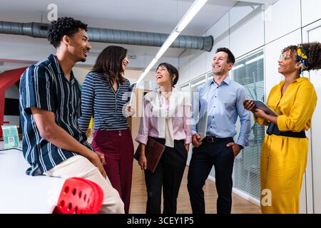 Five smiling business people are discussing together new company projects, walking through the office and holding notebooks, laptops and tablets Stock Photo