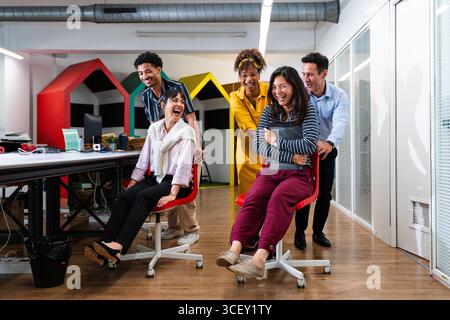 Multi-ethnic business team members are enjoying a playful moment, pushing each other on rolling chairs in a vibrant and modern office environment, fos Stock Photo