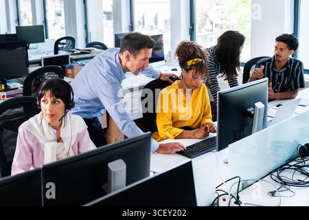 Office workers are collaborating on a project, using computers and discussing strategies in a busy office environment, showcasing teamwork and product Stock Photo