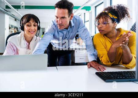 Business colleagues are actively participating in a collaborative project, engaging in a discussion around a laptop in a contemporary office setting, Stock Photo
