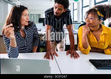 Three diverse colleagues are engaging in a productive meeting, analyzing data on a laptop and discussing strategies for their startup business in a mo Stock Photo