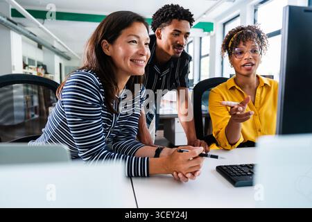 Three smiling business people are collaborating on a project, gathered around a desktop computer in a bright, modern office, discussing strategies and Stock Photo