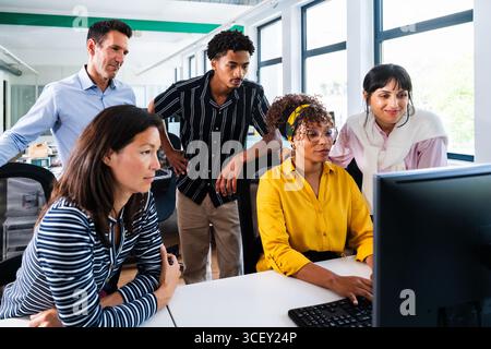 Diverse team of business professionals gathered around computer, analyzing data and discussing project in bright, modern office environment, showcasin Stock Photo