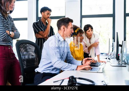Diverse colleagues are collaborating on a project, using laptops and desktop computers in a bright, modern office environment, discussing strategies a Stock Photo