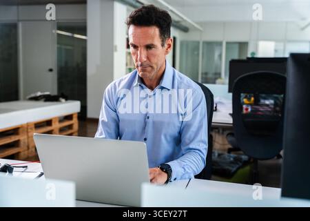 Concentrated mature businessman using laptop, sitting at desk in contemporary office, working on project, developing new ideas for startup company, de Stock Photo