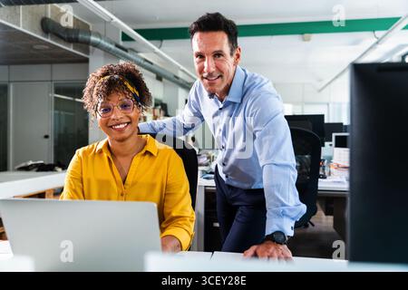 Two smiling business colleagues collaborating on a project while using a laptop in a modern, open-plan startup office, embodying teamwork and creativi Stock Photo