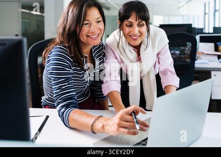Two smiling businesswomen are working together, analyzing data on a laptop in a bright, modern office, demonstrating teamwork and collaboration Stock Photo