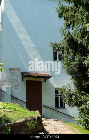 Old Wooden Door with Weathered Walls and Iron-Barred Window, Framed by Pine Tree and Stone Path Stock Photo