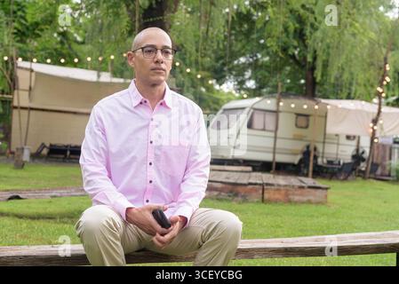 Bald Hispanic mature male in 40s wearing pink shirt and eyeglasses outdoors with camping park environment and caravan background. Concept of travel, t Stock Photo