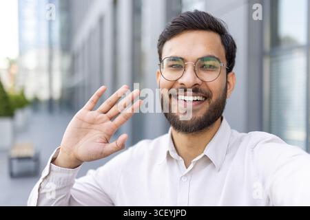 A cheerful person wearing glasses and a beard waves while smiling in an urban setting, conveying happiness and friendliness. Stock Photo