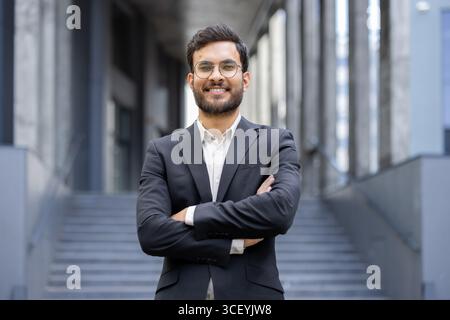 Professional young businessman smiling confidently while standing in an urban setting with arms crossed, showcasing success and ambition. The individual is wearing formal business attire Stock Photo
