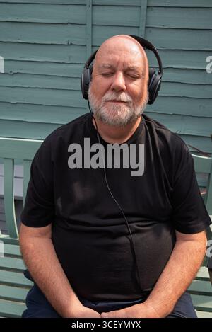 Senior man relaxing with headphones on a bench, eyes closed. Enjoying music or meditation. Stock Photo
