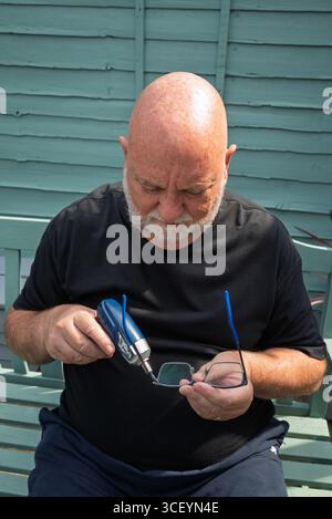 Man repairing glasses with a small screwdriver. DIY eyewear repair. Old man on smart phone with coffee. senior man and technology in use Stock Photo
