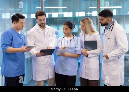 Medical staff collaborating to review patient information and discuss treatment plans. The group demonstrates teamwork, focusing on solutions. They are equipped with modern devices in a professional healthcare environment. Stock Photo