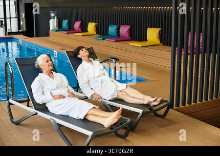 Two beautiful mature women unwind in spa robes beside a serene pool area, embracing tranquility. Stock Photo