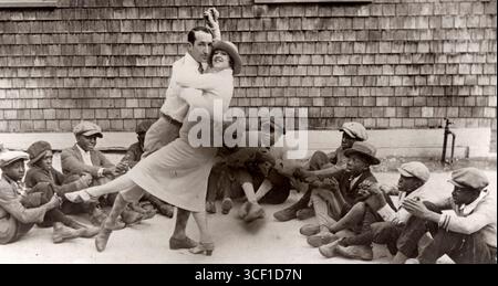 A man and woman performing the 'Charleston,' a dance associated with the Jazz Age. The performance takes place in the United States, 1926, with Black boys clapping in rhythm around them. Stock Photo