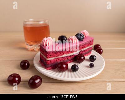Slice of berry cake with pink frosting, topped with cherries and blueberries, on a white plate. A pink-orange drink in the background adds charm. Grea Stock Photo