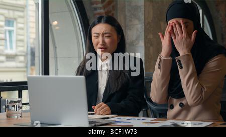 Two multiethnic business women in office looking computer screen shocked emotion negative amazed Asian woman Muslim Islamic girl diversity females Stock Photo