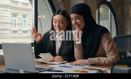 Two multiracial business women females happy talking video call corporate meeting negotiate writing notes analyze documents Asian Muslim businesswomen Stock Photo