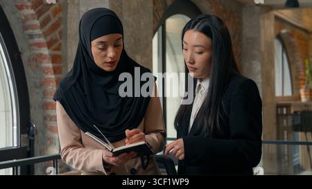 Serious concentrated two business women discussing startup project writing notes in notebook organizer planning job strategy multiracial females Stock Photo