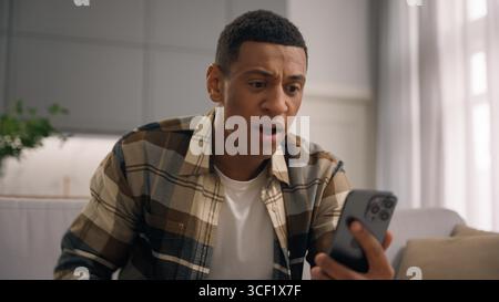 Furious negative mad African American man male guy irritated talking mobile phone chat virtual conference video call smartphone at home couch upset Stock Photo