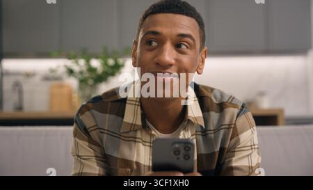 Joyful smiling African American man male guy at home using mobile phone looking for idea thinking carefree browsing smartphone application device Stock Photo