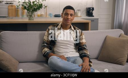 Portrait at home smiling cheerful candid male guy young calm resting ethnic African American man in living room looking at camera at couch alone Stock Photo