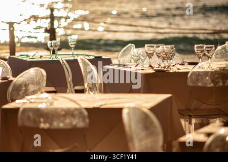 Elegant restaurant tables are set for dinner, overlooking a sparkling ocean at sunset, creating a romantic and luxurious dining experience Stock Photo