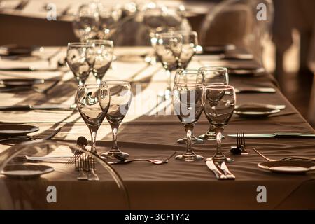 Empty wine glasses and cutlery are placed on a long table in a restaurant, creating an elegant setting for an upcoming event at sunset Stock Photo