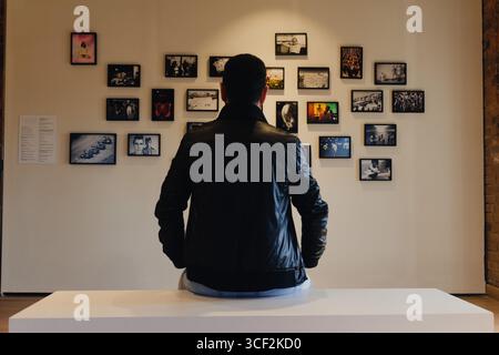 Rear view of a man in a black leather jacket sitting on a bench and looking at a wall of framed photographs in a minimalist art gallery Stock Photo