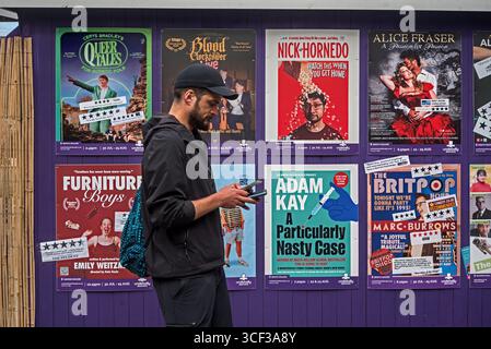 Man looking at his phone walking by a wall of Edinburgh Fringe Show posters, Edinburgh, Scotland, UK. Stock Photo