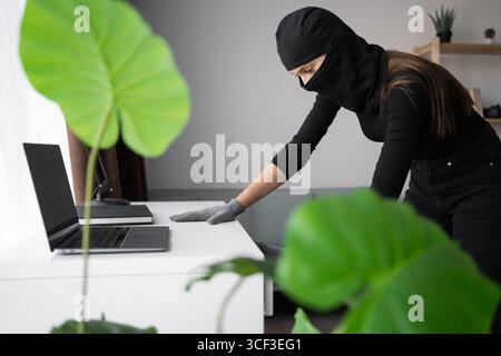 Female thief or burglar in black balaclava stealing home standing near table with laptop computer. Theft Stock Photo