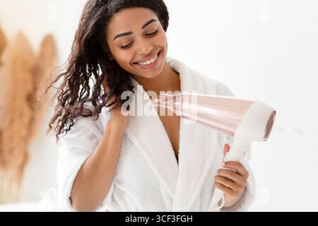 Happy Black Lady Drying Wavy Hair With Hairdryer In Bathroom Stock Photo