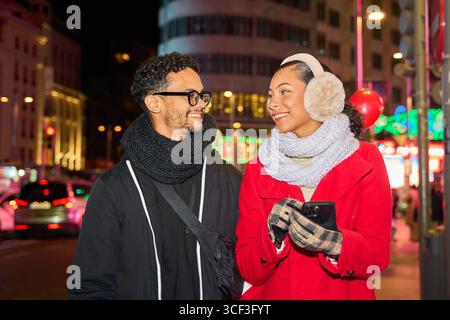 Young loving couple walking in a decorated city street at night during christmas time, looking at each other and using a smartphone Stock Photo