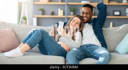 Cheerful multicultural spouses relaxing on couch with smartphone together Stock Photo