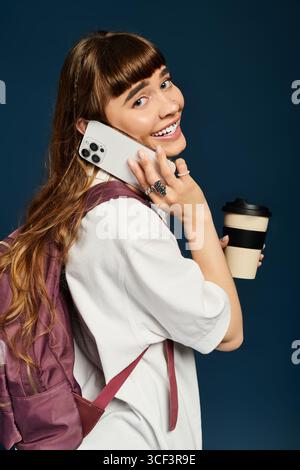 A young woman with a joyful smile talks on her phone, holding a coffee cup, ready for her studies. Stock Photo