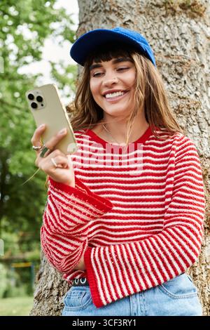 A young woman in a striped sweater smiles at her phone, leaning against a tree in the park. Stock Photo