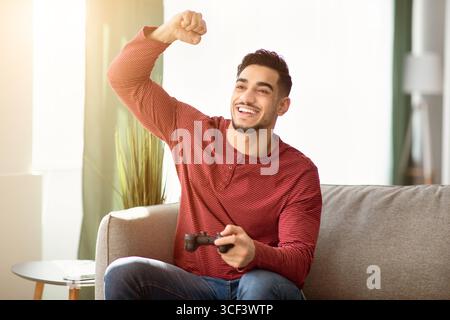 Happy arab man sitting on couch in living room, gaming Stock Photo