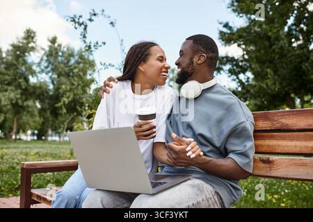 A cheerful couple shares laughter while using a laptop on a park bench, surrounded by greenery. Stock Photo
