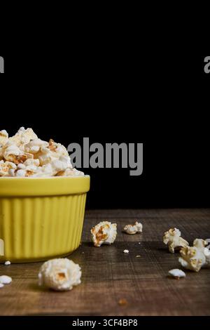 Bowl of popped corn on a wooden surface, with a black background that highlights color and texture. Ideal for editorial, gastronomic, or advertising Stock Photo