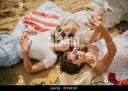 Three women are laying on the beach, one of them taking a selfie with her cell phone. Scene is relaxed and carefree, as the women are enjoying their t Stock Photo