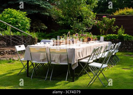A long dining table set for an outdoor gathering, surrounded by greenery. the table is elegantly arranged with centerpieces, offering a peaceful al fr Stock Photo