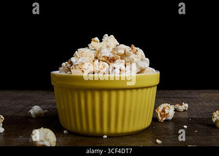 Yellow bowl with popcorn on wood, deep black background. Centered composition, with less dispersion. Ideal for product photography or minimalist food Stock Photo