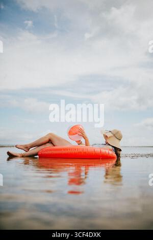 A woman is floating on a red inflatable raft in the ocean. She is holding a pink book in her lap. The scene is peaceful and relaxing, with the woman e Stock Photo
