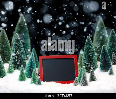Blank chalkboard with copy space for Christmas message surrounded by miniature trees and falling snow. Stock Photo