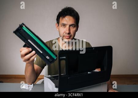 Studio portrait of computer technician replacing toner cartridge in laser printer during maintenance service, holding new cartridge and looking at Stock Photo