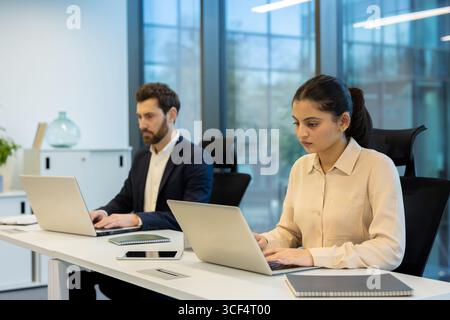 A man and woman concentrating on tasks while using laptops in a contemporary office. Bright atmosphere with sleek furnishings enhances the professional setting and signifies productivity Stock Photo