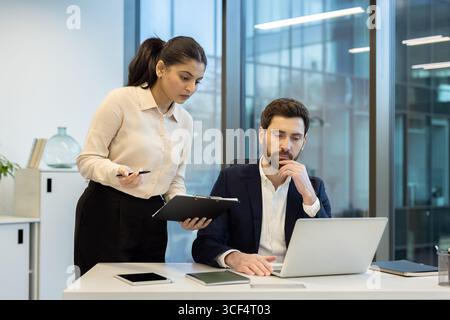 A professional scene showing two colleagues working together in a modern office. One is seated at a desk using a laptop, while the other provides assistance and references notes on a clipboard. Stock Photo